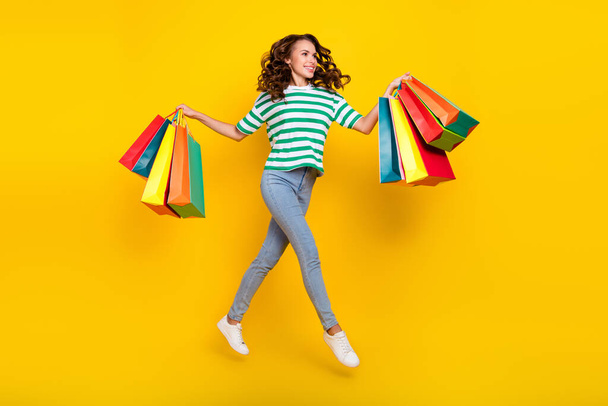 Full body photo of impressed brunette young lady run with bags wear t-shirt jeans footwear isolated on yellow background. - Valokuva, kuva