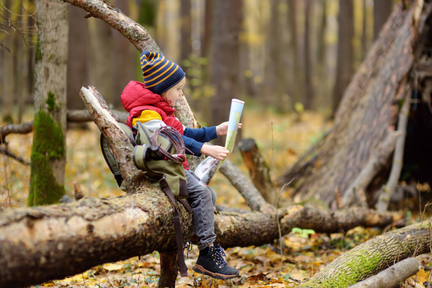 Little boy scout is orienteering in forest. Child is sitting on fallen tree and looking on map on background of teepee hut. Concepts of adventure, scouting and hiking tourism for kids. - Foto, Imagem