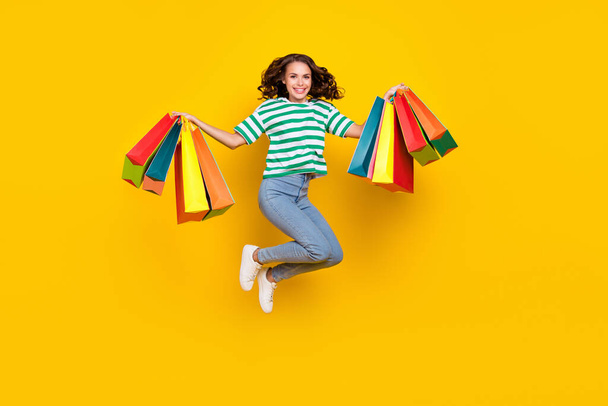 Full length photo of cool brunette millennial lady jump with bags wear t-shirt jeans shoes isolated on yellow background. - Valokuva, kuva