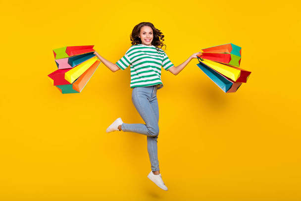 Full body photo of impressed brunette young lady run with bags wear t-shirt jeans sneakers isolated on yellow background. - Foto, immagini
