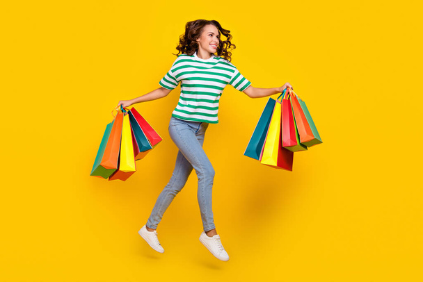 Full length photo of cute brunette millennial lady run with bags wear t-shirt jeans footwear isolated on yellow background. - Zdjęcie, obraz