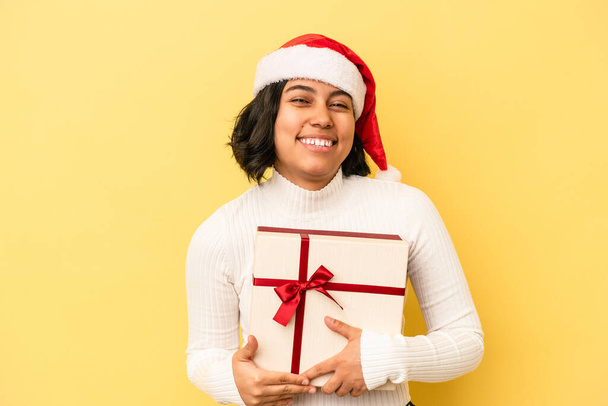 Young latin woman celebrating christmas holding a gift isolated on yellow background laughs out loudly keeping hand on chest. - Фото, изображение