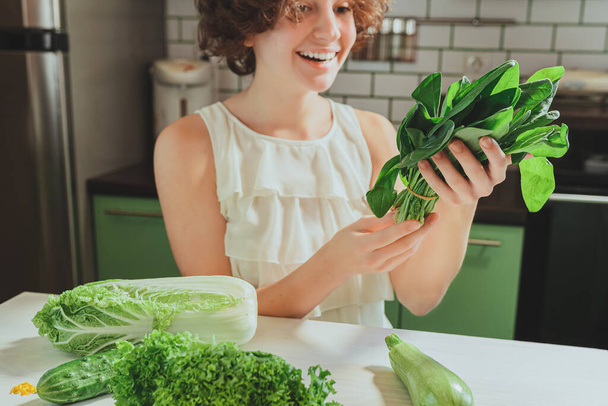 smiling girl with brown curly hair, in white blouse, standing in home kitchen, holds freshly picked spinach greens in hands, rejoices in buying fresh vegetables, preparing vitamin salad or cocktail. green vegetables and herbs - Zdjęcie, obraz