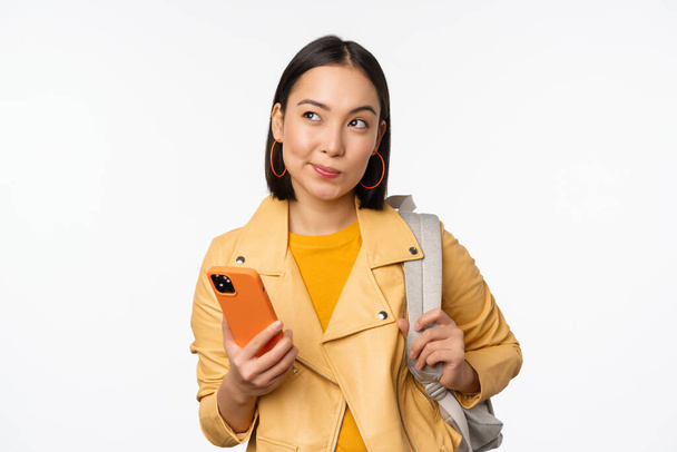 Asiatique fille voyageur avec sac à dos, tenant téléphone mobile, en utilisant l'application smartphone, regardant réfléchi, debout sur fond blanc - Photo, image