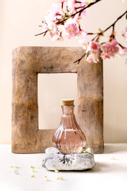 Glass pink transparent bottle of perfume with cork, standing on textured stone on white marble table with white flowers and blossom spring branch. Perfume making concept with place for design label - 写真・画像