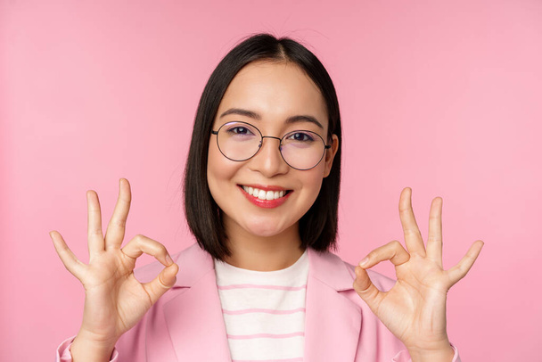 Detail portrét ohromen korporátní žena, asijské obchodní dáma v brýlích, ukazující v pořádku znamení, při pohledu na kameru, doporučující, růžové pozadí - Fotografie, Obrázek