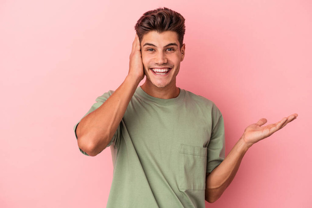 Young caucasian man isolated on pink background holds copy space on a palm, keep hand over cheek. Amazed and delighted. - Photo, Image