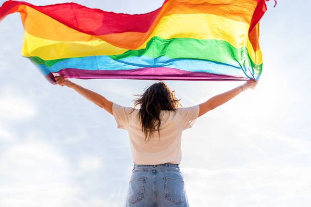 Νεαρή γυναίκα κυματίζει σημαία ΛΟΑΤΚΙ κοιτάζοντας τον ήλιο. - Φωτογραφία, εικόνα
