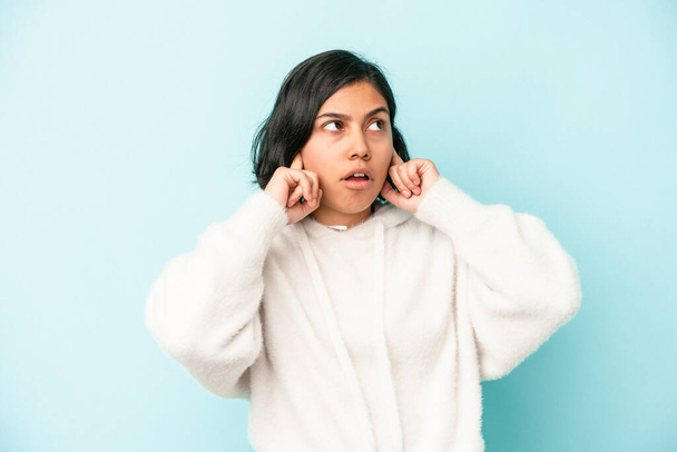 Mujer latina joven aislada sobre fondo azul cubriendo las orejas con los dedos, estresada y desesperada por un ambiente ruidoso. - Foto, Imagen