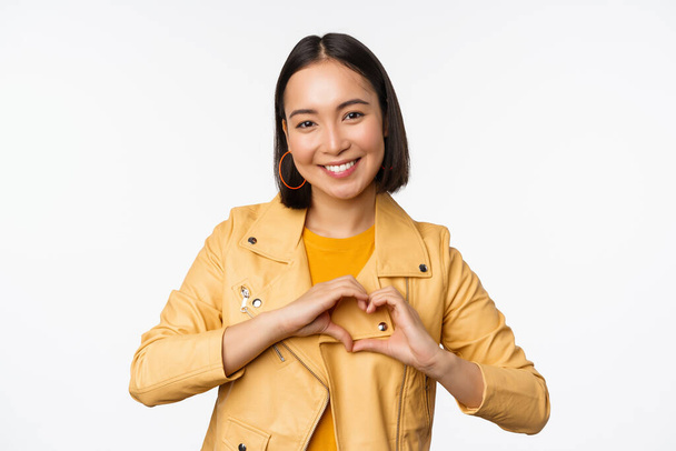 Image of beautiful asian girl showing heart, love sign, express care and romantic feelings, standing over white studio background - Zdjęcie, obraz