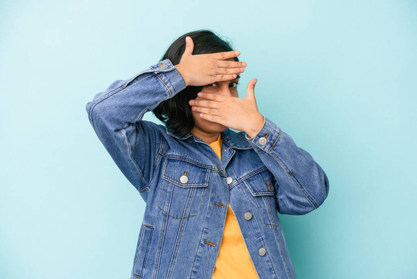 jong latijn vrouw geïsoleerd op blauwe achtergrond knipperen op de camera door vingers, beschaamd bedekking gezicht. - Foto, afbeelding
