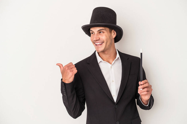 Young magician caucasian man holding a wand isolated on white background points with thumb finger away, laughing and carefree. - Photo, Image