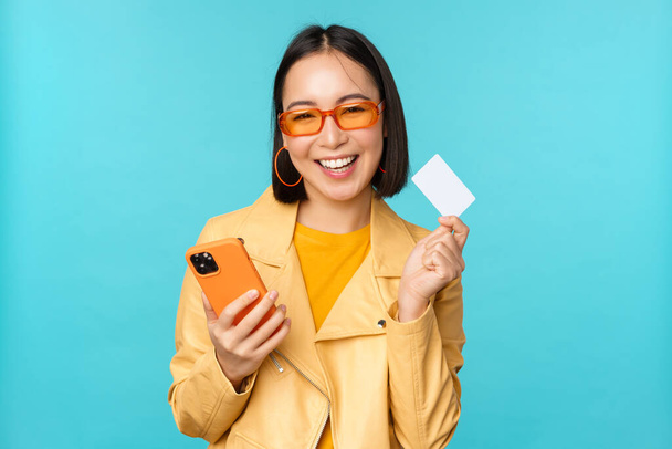 Online shopping. Stylish young asian woman in sunglasses, showing credit card and using smartphone, paying in internet, making purchase, standing over blue background - Photo, Image