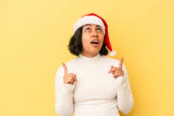 Młoda latynoska kobieta świętująca Boże Narodzenie odizolowana na żółtym tle wskazując do góry nogami z otwartymi ustami. - Zdjęcie, obraz