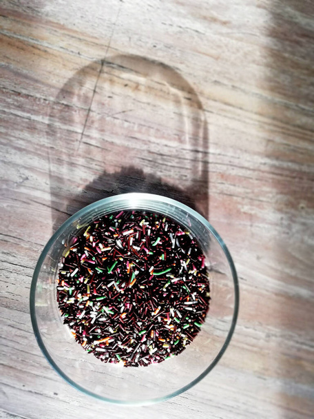 Granulki czekoladowe o wielu kolorach, umieszczone na szklanej misce, z drewnianym tłem stołu i cieniem - Zdjęcie, obraz