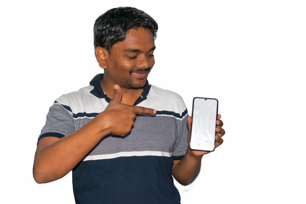 Opgewonden knappe Indiase man met mobiele telefoon, wijzend met de vinger naar het scherm en glimlachend, staande tegen een witte achtergrond. Wit scherm voor kopieerruimte. - Foto, afbeelding