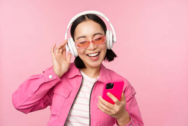 Stijlvol Aziatisch meisje dansen met smartphone, muziek luisteren in koptelefoon op mobiele telefoon app, glimlachen en lachen, poseren tegen een roze achtergrond - Foto, afbeelding