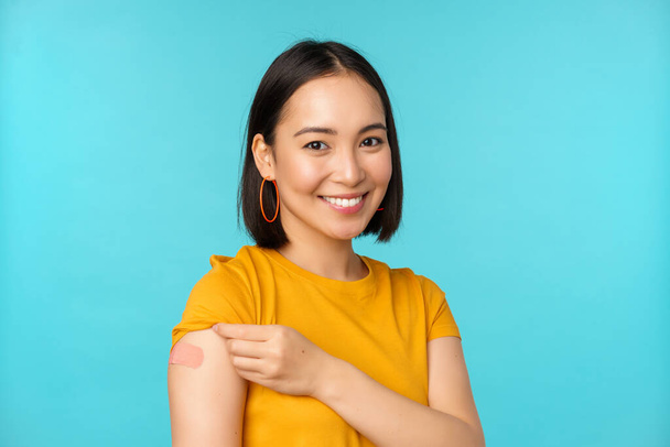 Campaña de vacunas de covid-19. Joven hermosa, saludable mujer asiática mostrando hombro con bandaid, concepto de vacunación, de pie sobre fondo azul - Foto, imagen