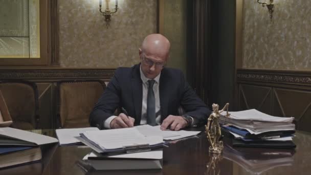Προσωπογραφία μεσήλικου δικηγόρου με επίσημο κοστούμι και γυαλιά που γράφει σε χαρτί και ποζάρει για κάμερα στο χώρο εργασίας στο γραφείο - Πλάνα, βίντεο