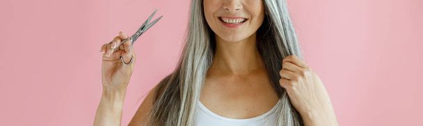 Cheerful Asian lady holds lock of silver hair and scissors on pink background - Foto, immagini