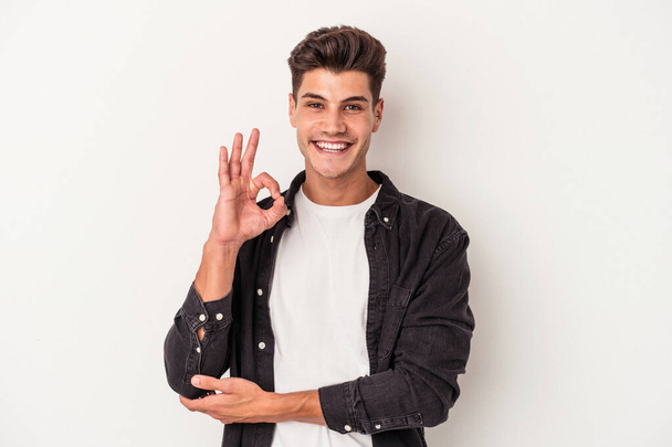 Young caucasian man isolated on white background winks an eye and holds an okay gesture with hand. - Photo, Image