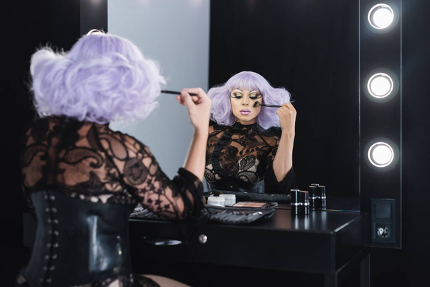 man in purple wig applying makeup near mirror in dressing room - Photo, Image