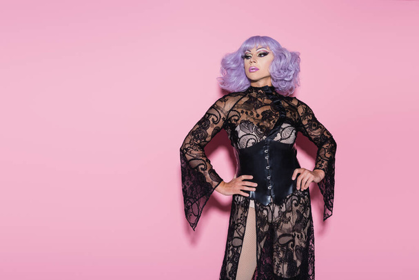 drag queen με φόρεμα και βιολετί περούκα που ποζάρει με τα χέρια στους γοφούς και κοιτάζει μακριά στο ροζ - Φωτογραφία, εικόνα