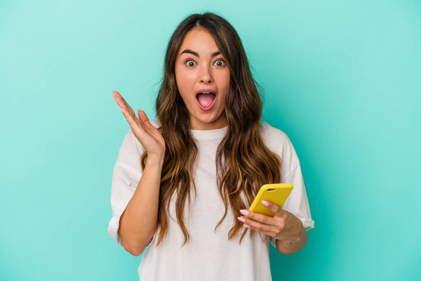 Молодая белая женщина с мобильным телефоном на синем фоне удивлена и шокирована. - Фото, изображение