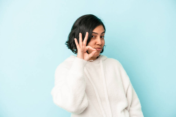 Young latin woman isolated on blue background winks an eye and holds an okay gesture with hand. - Photo, Image