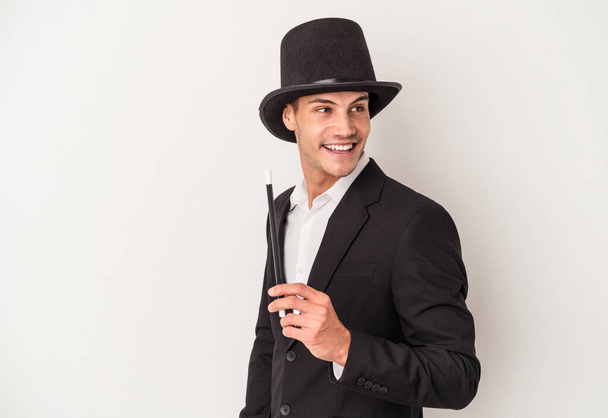 Young magician caucasian man holding a wand isolated on white background looks aside smiling, cheerful and pleasant. - Photo, Image