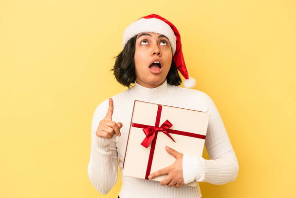 Молода латинка святкує Різдво, що тримає подарунок ізольовано на жовтому тлі, спрямованому догори з відкритим ротом
. - Фото, зображення