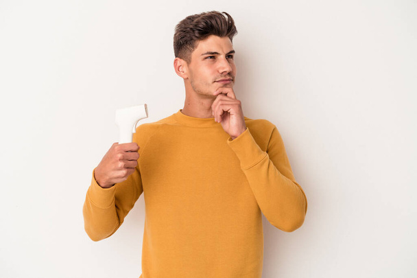 Homem caucasiano jovem segurando termômetro isolado no fundo branco olhando para os lados com expressão duvidosa e cética. - Foto, Imagem