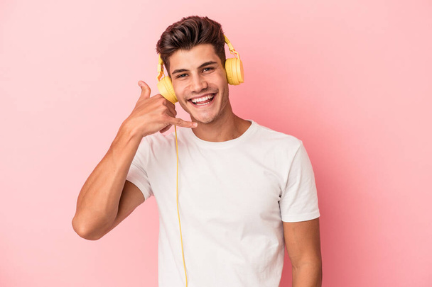 ピンクの背景に孤立した音楽を聴く若い白人男性指で携帯電話のコールジェスチャーを示しています. - 写真・画像