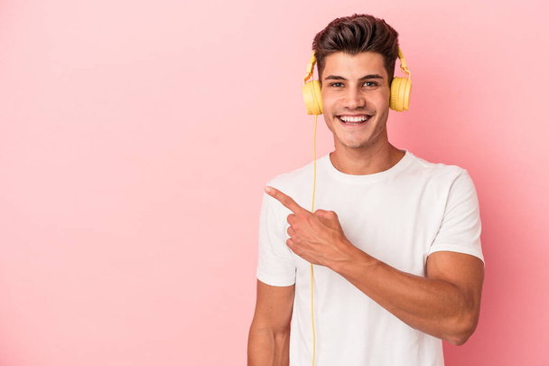 Giovane uomo caucasico che ascolta musica isolata su sfondo rosa sorridendo e indicando da parte, mostrando qualcosa nello spazio vuoto. - Foto, immagini