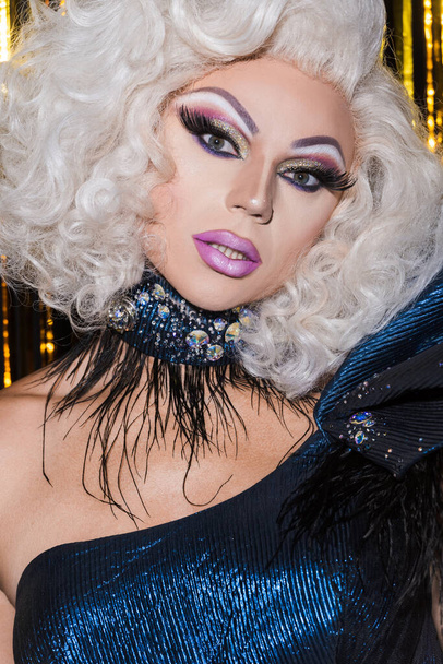 drag queen με εντυπωσιακό μακιγιάζ κοιτάζοντας κάμερα σε λαμπερό φόντο - Φωτογραφία, εικόνα