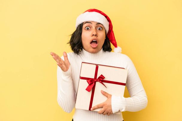 Молода латинка, яка святкує Різдво, тримає подарунок ізольовано на жовтому тлі, здивована та шокована
. - Фото, зображення