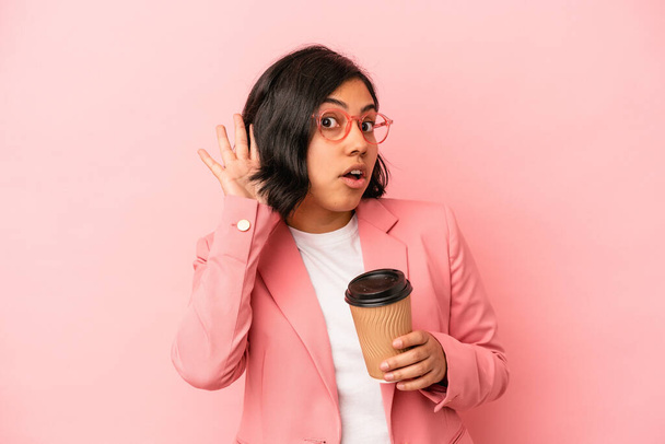 Молода латинка тримає каву ізольовано на рожевому фоні, намагаючись слухати плітки
. - Фото, зображення