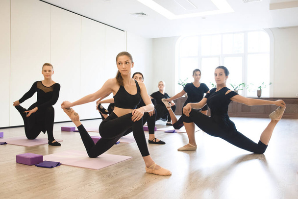 Active, healthy lifestyle, hobby, recreation, wellbeing, weight loss concepts. Group dancer girls warming up with stretching exercises for flexibility during class in white sports hall - Photo, Image