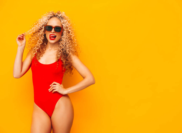 Young beautiful smiling woman posing near yellow wall in studio.Sexy model in red swimwear bathing suit.Positive female with curls hairstyle.Happy and cheerful. In sunglasses - Foto, Imagen