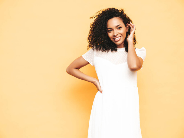 Portrait of beautiful black woman with afro curls hairstyle. Smiling model dressed in white summer dress. Sexy carefree female posing near yellow wall in studio. Tanned and cheerful - Foto, Bild