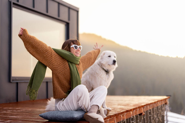 Жінка з собакою відпочиває на терасі крихітного будинку на природі
 - Фото, зображення