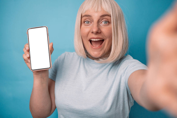 Happy young 20s woman showing empty cell phone screen on blue studio background. Excited lady recommending mobile application or website, offering space for your ad, mockup - Фото, изображение