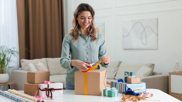 Beautiful young woman wrapping gift at table in living room - Photo, image