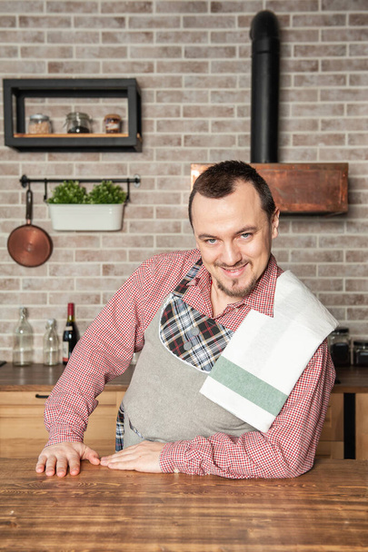 Engraçado bonito jovem sorrindo homem vestindo pinafore com toalha na cozinha - Foto, Imagem