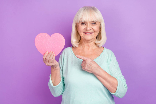 Photo of nice pensioner romantic lady direct finger pink heart figure wear teal shirt isolated violet color background - 写真・画像