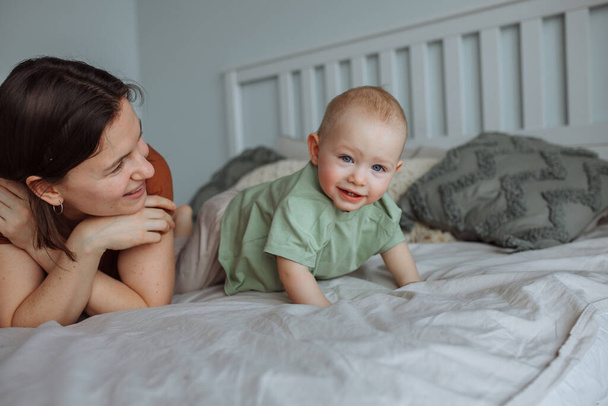 A young brunette mother in a brown shirt has fun at home with her one-year-old son on the bed. Scandinavian interior style - Fotó, kép