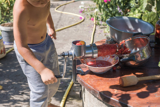 Hausgemachter Tomatensaft wird in einer Handpresse mit einer Schraube gepresst Kinder helfen, den Stielsaft aus dem Fruchtfleisch durch ein Sieb zu drehen, das Fruchtfleisch wird auf den Kompost geworfen - Foto, Bild