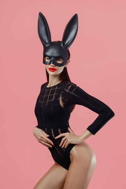Sexy woman wearing a black mask Easter bunny standing on a pink background and looks very sensually. - Zdjęcie, obraz