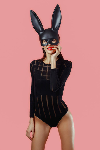 Sexy woman wearing a black mask Easter bunny standing on a pink background and looks very sensually. - Photo, Image