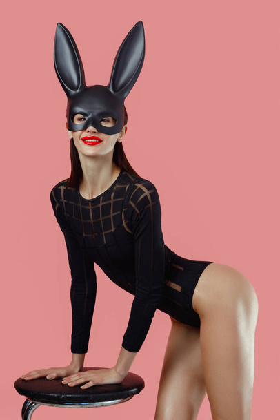 Siyah maskeli seksi bir kadın. Paskalya tavşanı pembe arka planda duruyor ve çok şehvetli görünüyor.. - Fotoğraf, Görsel